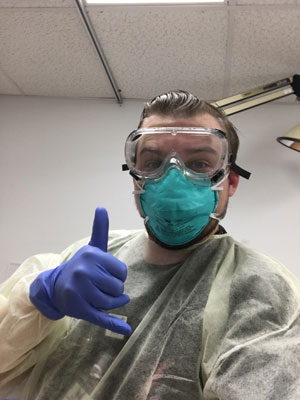 Picture of a male Nurse in the Emergency Care Department wearing goggles, a face mask, gloves, and giving a "hang loose" sign with his hand.